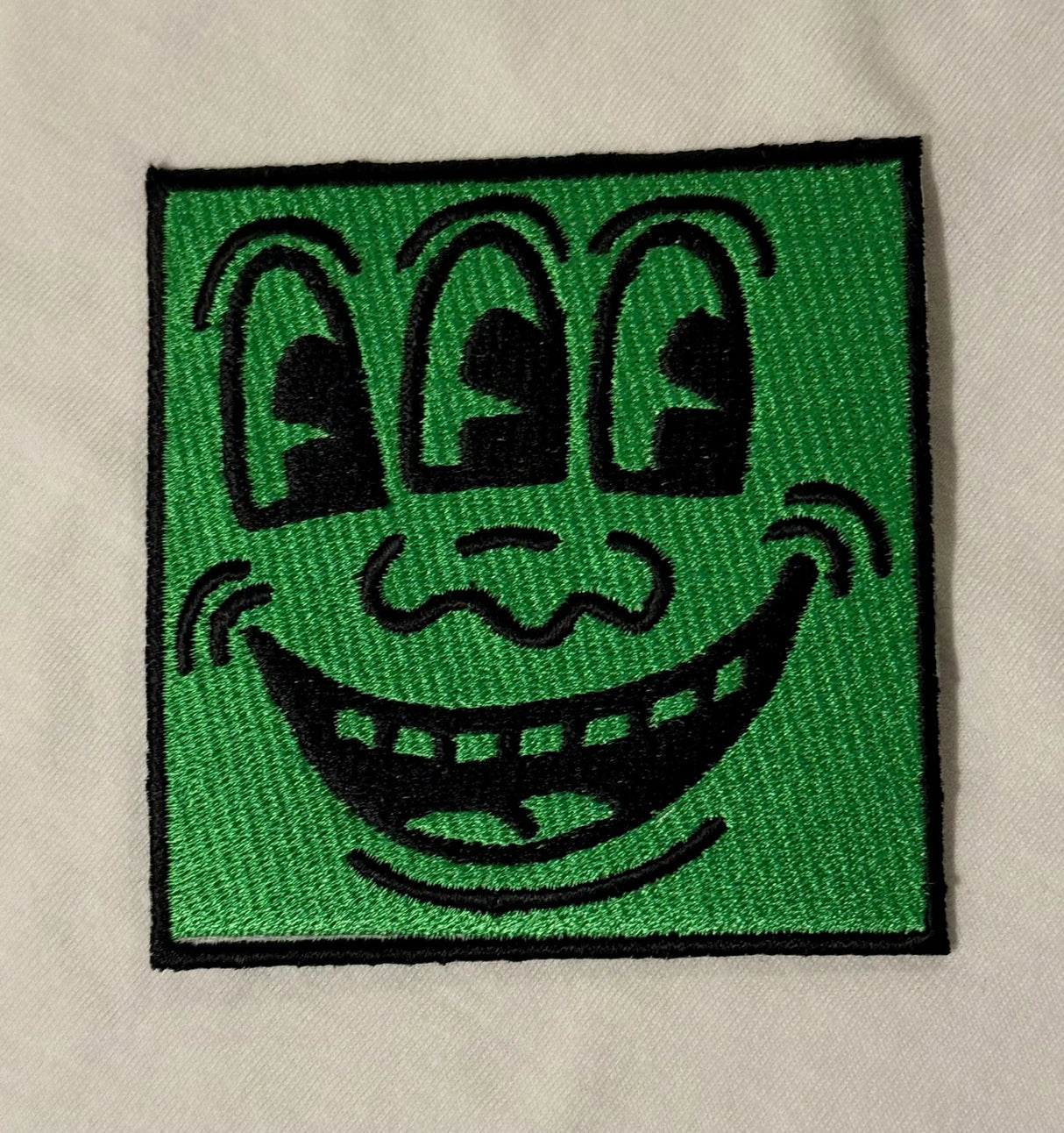 Embroidered patch with a cartoonish face design on a green background, attached to a beige fabric.