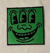 Embroidered patch with a cartoonish face design on a green background, attached to a beige fabric.