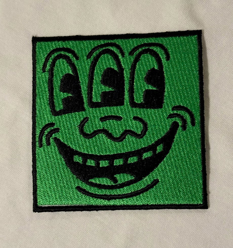 Embroidered patch with a cartoonish face design on a green background, attached to a beige fabric.