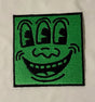 Embroidered patch with a cartoonish face design on a green background, attached to a beige fabric.