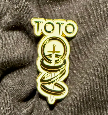 Gold Toto band logo pin on a dark fabric background