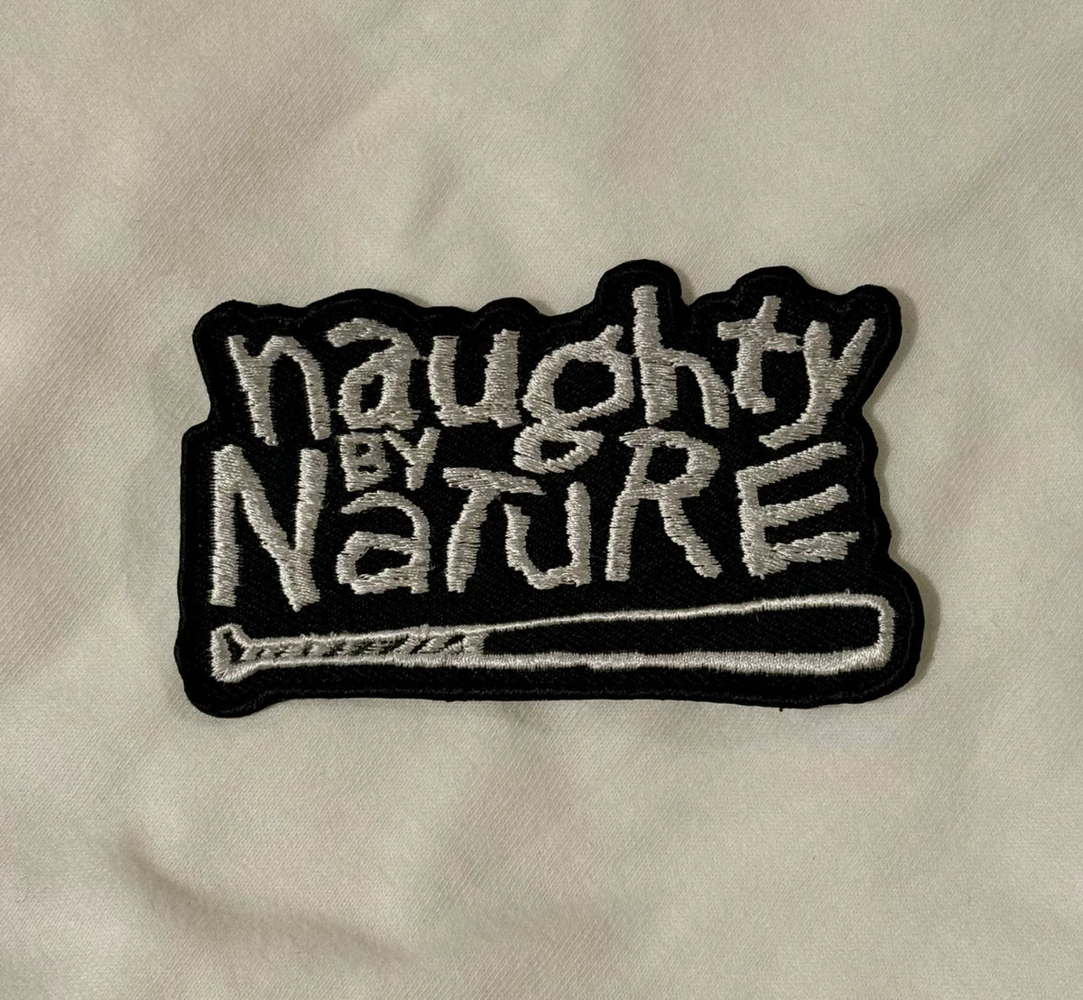 An embroidered patch with the text 'naughty BY NATURE' in white thread on a black background, featuring a baseball bat graphic at the bottom, placed on a light fabric surface.