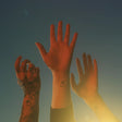 Three hands raised in the air against a blue sky background, symbolizing unity or a communal spirit, possibly related to a music album or event.