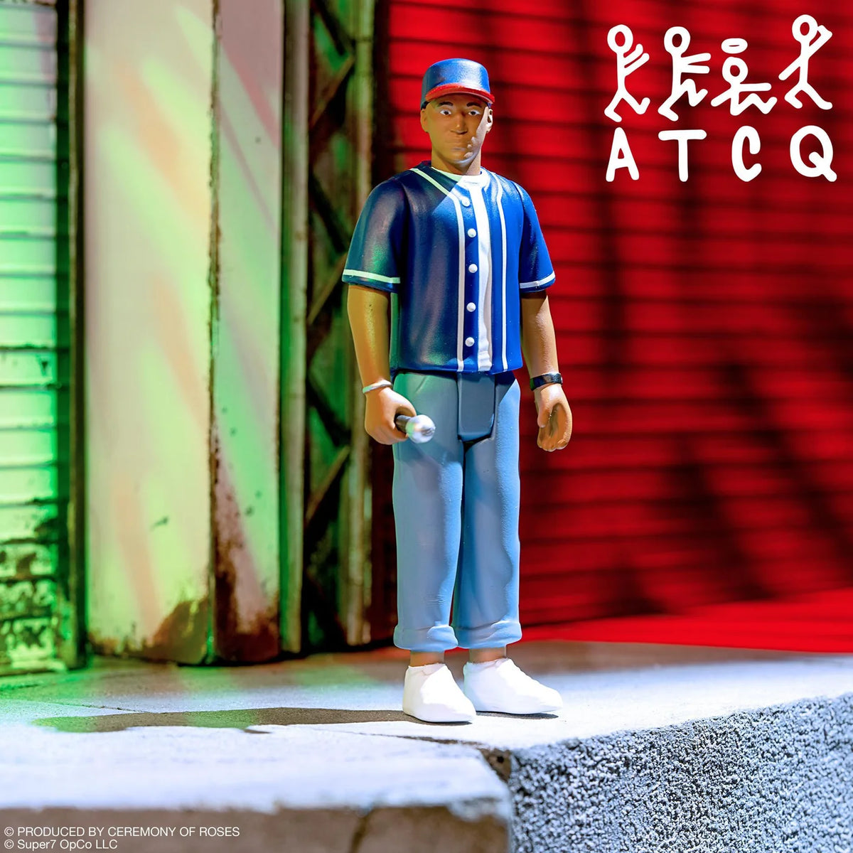 A collectible action figure of a baseball player wearing a blue and white striped jersey, blue pants, and a blue cap, standing on a concrete platform with a red and green striped background. The text 'ATCQ' is visible in the top right corner.