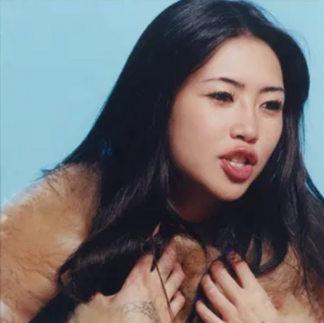 This image features the artist beabadoobee, portrayed with a playful expression, sticking out her tongue. She is dressed in a plush, furry garment, adding an edgy yet charming vibe to the photo.