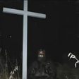A person wearing a mask sitting in front of a cross in a dark environment, possibly as a symbolic or artistic representation related to the album's themes.