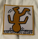 An embroidered square patch featuring a stylized upside-down figure in mustard yellow with black outlines on a white background, bordered in yellow, placed on a white fabric surface.