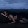 Woman lying on a dark surface with a mountainous landscape under a cloudy sky.