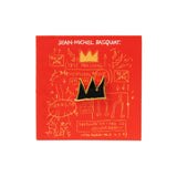 A black and gold crown-shaped enamel pin by Jean-Michel Basquiat, mounted on a red backing card with gold graffiti-style drawings and text.