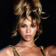 Beyonce Knowles in a high-volume messy bun hairstyle with blonde and brown highlights, wearing a gold chain-strap top against a solid black background.