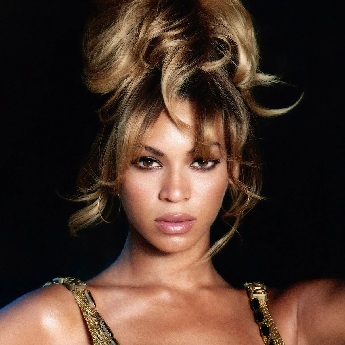 Beyonce Knowles in a high-volume messy bun hairstyle with blonde and brown highlights, wearing a gold chain-strap top against a solid black background.