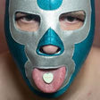 Person wearing a blue and silver mask with a heart-shaped candy on their tongue.