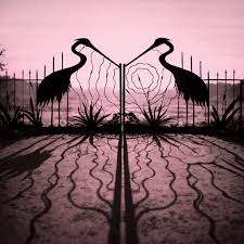 A decorative metal gate with two large crane silhouettes and a central spiral motif, shown in a symmetrical garden setting with a pinkish-purple sky and strong shadows.