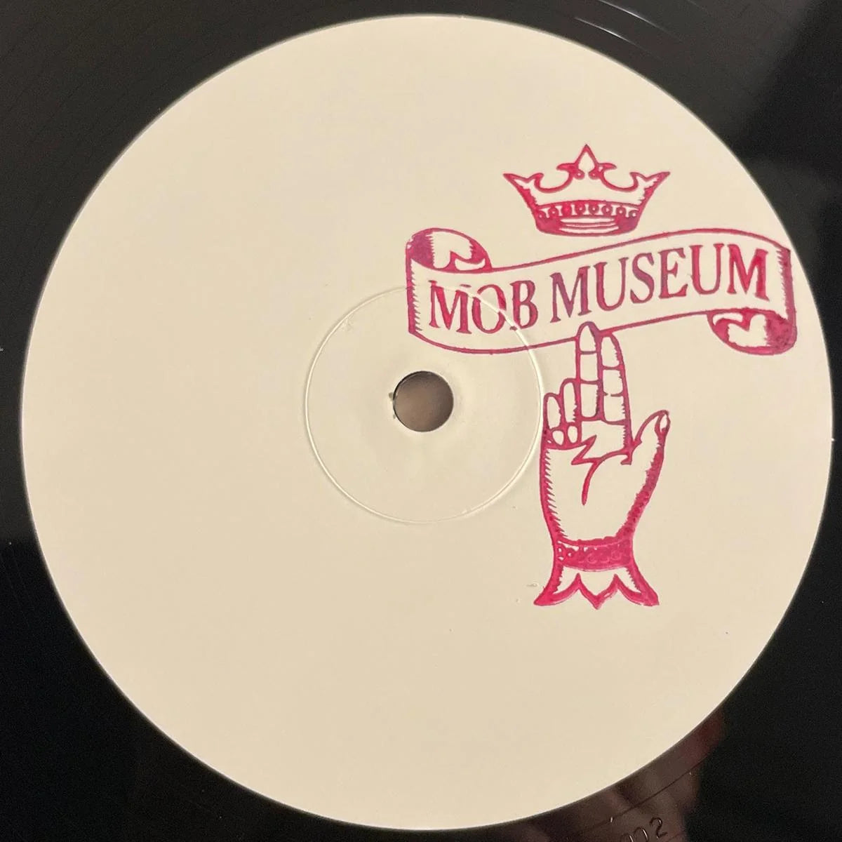 A close-up of a vinyl record label featuring a pink graphic of a hand holding a banner that reads 'MOB MUSEUM' under a crown, on a black vinyl disc.