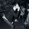 Black and white photo of a person singing into a microphone on a stage, wearing a dark textured sweater and jeans, with a crowd and graffiti in the background.