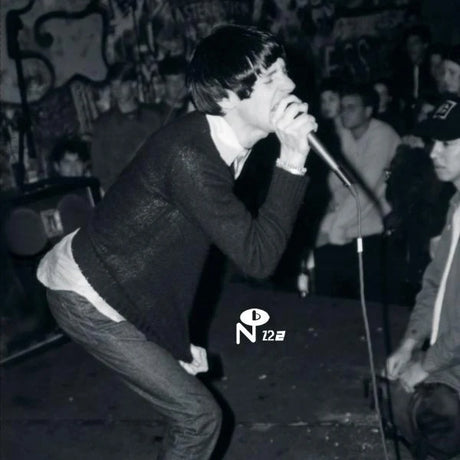 Black and white photo of a person singing into a microphone on a stage, wearing a dark textured sweater and jeans, with a crowd and graffiti in the background.