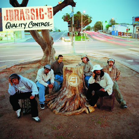 The image shows the members of Jurassic 5 sitting around a large tree stump with the album 'Quality Control' written above. They are dressed in casual clothing and the setting appears to be an outdoor location, possibly a street or park.
